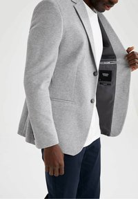 Blazer gris en una tela texturizada, de un solo botón con dos botones, presenta bolsillos laterales y un forro negro con una etiqueta en el interior.