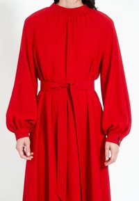 Červené dlouhé šaty s dlouhým rukávem a vysokým výstřihem, s nařasenými detaily, páskem v oblasti pasu a objemnými rukávy. Hladká látka s plisovými akcenty.