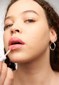 Lipgloss-applicator dicht bij de lippen gehouden. De glans heeft een zachte roze tint en een gladde textuur. Het model heeft een zichtbare neus en draagt ringoorbellen.