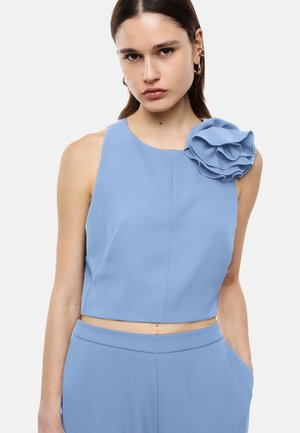 Donna che indossa un top corto senza maniche color azzurro chiaro con un grande fiore in tessuto sulla spalla e pantaloni abbinati a vita alta, in piedi con una mano in tasca.