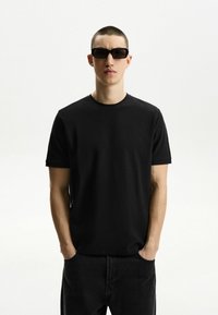 Schwarzes Rundhals-T-Shirt aus strukturiertem Stoff, kombiniert mit dunklen Denim-Hosen. Eine rechteckige Sonnenbrille rundet das Outfit ab.
