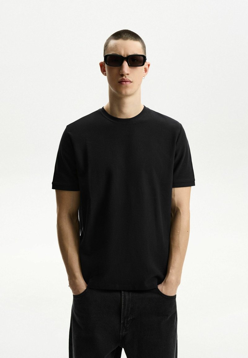 Schwarzes Rundhals-T-Shirt aus strukturiertem Stoff, kombiniert mit dunklen Denim-Hosen. Eine rechteckige Sonnenbrille rundet das Outfit ab.