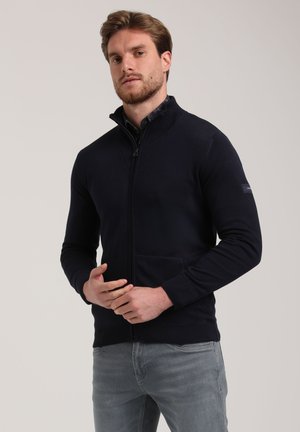 Suéter navy con cierre, cuello acanalado, dos bolsillos frontales y patrón de punto texturizado. Lleva encima de una camisa de cuello, con silueta ajustada.