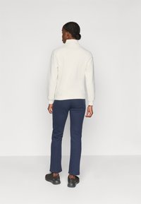 Sudadera cremosa con cremallera y cuello alto, combinada con pantalones azul marino y zapatillas negras, vista desde atrás. Textura suave, corte relajado.