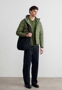 Νεαρός άνδρας που φοράει πράσινο μπουφάν με φουσκωτά μέρη, πράσινο πλεκτό πουκάμισο, μαύρο παντελόνι, μαύρα παπούτσια, κρατώντας μια μεγάλη μαύρη τσάντα ώμου σε εσωτερικό χώρο.