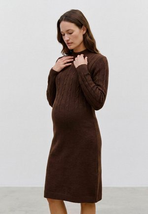 Femme enceinte portant une robe marron en maille torsadée à manches longues, touchant son col, debout devant un fond gris clair uni.