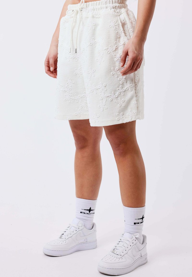 Personne portant un short blanc à motifs floraux texturés avec cordon de serrage, des chaussettes blanches montantes avec logo noir, et des baskets blanches, debout sur un fond uni.