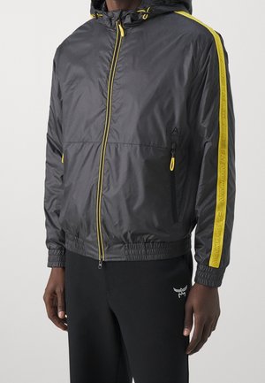 Mężczyzna w czarnej kurtce z kapturem z żółtym zamkiem błyskawicznym i żółtym paskiem na rękawach z logo "EA7 Emporio Armani".