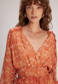 Orangenes Kleid mit V-Ausschnitt, langen Ärmeln und gerafftem Taille. Verfügt über ein gewelltes Muster und Rüschendetails an den Schultern.