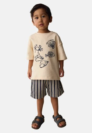 Jonge jongen draagt een beige oversized T-shirt met prints van zeedieren, gestreepte korte broek en donkere sandalen, staand tegen een effen achtergrond.