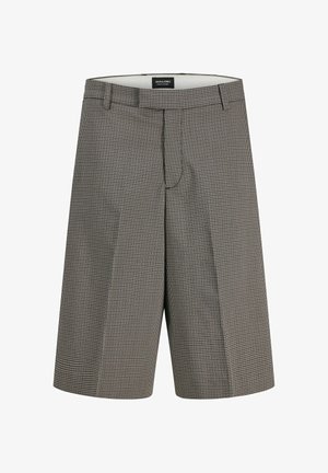 Garçon aux cheveux bouclés portant une veste noire, un pull en maille blanche, un short à motifs jusqu'aux genoux, des chaussettes blanches et des chaussures noires, debout les mains dans les poches.