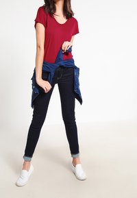 Kvinde iført en rød kortærmet skjorte, mørke stramme jeans med opfoldede ben, hvide slip-on sko og en blå jakke knyttet omkring taljen.