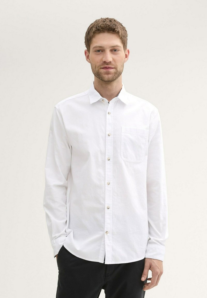 Chemise blanche à manches longues avec boutons en tissu léger, comportant une poche poitrine, une texture subtile et des boutons en forme de perle.