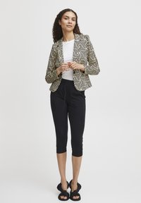 Blazer à motifs noir et crème avec un devant ouvert, associé à un t-shirt blanc et un legging noir cropped. Des sandales noires complètent le look.