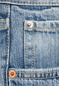 Gros plan sur un jean en denim bleu montrant les détails de la poche avec une couture jaune et des rivets métalliques sur les bords de la poche.