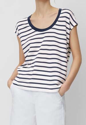 Femme portant un haut blanc sans manches avec des rayures horizontales bleu marine et un bord bleu marine, associé à un pantalon blanc, mains dans les poches.