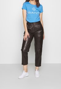 Blå bomullströja med GANT-logga, kombinerad med högmidjade, bruna läderbyxor och vita sneakers. Enkelt, åtsittande snitt.