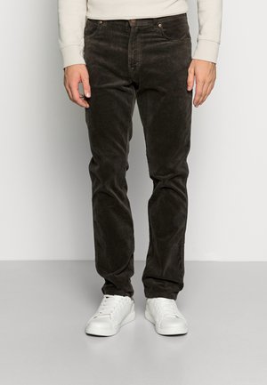 Mężczyzna w czarnych spodniach sztruksowych i białych trampkach, stojący na jasnoszarej podłodze przy gładkiej ścianie.
