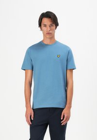 Tânăr în picioare, îmbrăcat cu un tricou simplu albastru cu un mic logo de pasăre galbenă pe pieptul stâng, asortat cu pantaloni închiși la culoare, fundal alb.