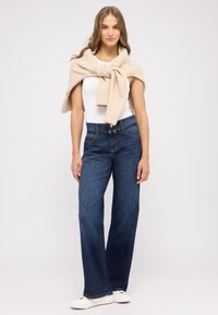 Weite blaue Jeans mit hoher Taille, kombiniert mit einem cremefarbenen Strickpullover, der über die Schultern gelegt ist, und weißen Schuhen.