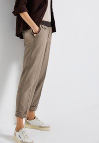 Pantalons marron clair en tissu tissé, présentant une coupe décontractée, des ourlets retroussés et des poches avant. Assortis à des baskets blanches et beige.
