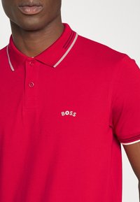 Röd polo-shirt i bomull med krage som har gråa och vita ränder. Inkluderar en liten silverfärgad "BOSS"-logotyp på vänster bröst.