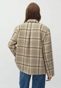 Beige kariertes Blazer mit schwarzen und orangefarbenen Akzenten, strukturiertem Design, langen Ärmeln und seitlichen Taschen. Kombiniert mit blauen Jeans.