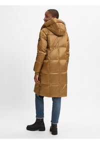 Bruine poffermantel met een capuchon, heeft gewatteerde delen, een logo-patch op de arm en een zijrits. Gepaard met blauwe jeans en zwarte laarzen.