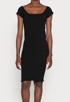 Personne portant une robe noire ajustée, côtelée, à manches courtes et à encolure carrée, se tenant devant un fond clair uni.