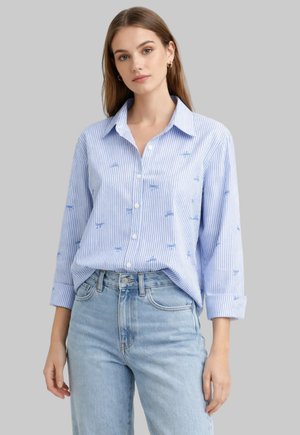 Giovane donna con capelli castani lisci indossa una camicia a righe azzurro chiaro con piccoli ricami blu e jeans azzurri.
