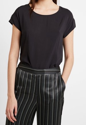 Femme portant un chemisier noir à manches courtes rentré dans un pantalon noir taille haute à rayures blanches verticales.
