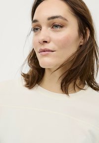 Lichtbeige top met een ronde hals, voorzien van subtiele stikselaccenten. Vlotte textuur en een ontspannen pasvorm, gedeeltelijk zichtbaar in het kader.
