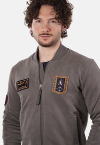 Giacca bomber grigia con zip frontale, colletto a coste e due tasche laterali, adornata da toppe tra cui "LEADER" e un'insegna.