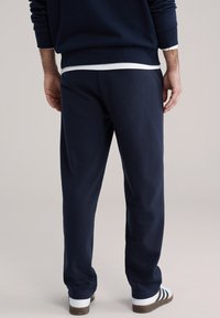 Mornarskoplave trenirka hlače opuštenog kroja, glatke teksture i s elastičnim pojasom. Uparene s bijelim tenisicama s crnim prugama.