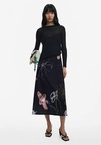 Ilgas juodas sijonas su spalvingomis gėlių raštais; derinamas su aptemptu juodu permatomu megztiniu ir laikomas struktūruotas rankinė.