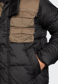 Il giubbotto puffo nero e marrone presenta molteplici tasche frontali, un tessuto testurizzato, colori a contrasto e un design del colletto aderente.