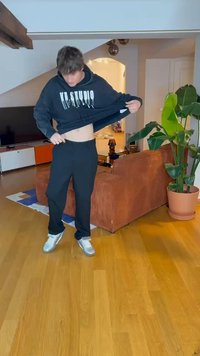 Sweatshirt preto com o texto "KL STUDIO", ajuste descontraído, usado com calças de moletom pretas e ténis prateados. O fundo inclui uma planta e um sofá.