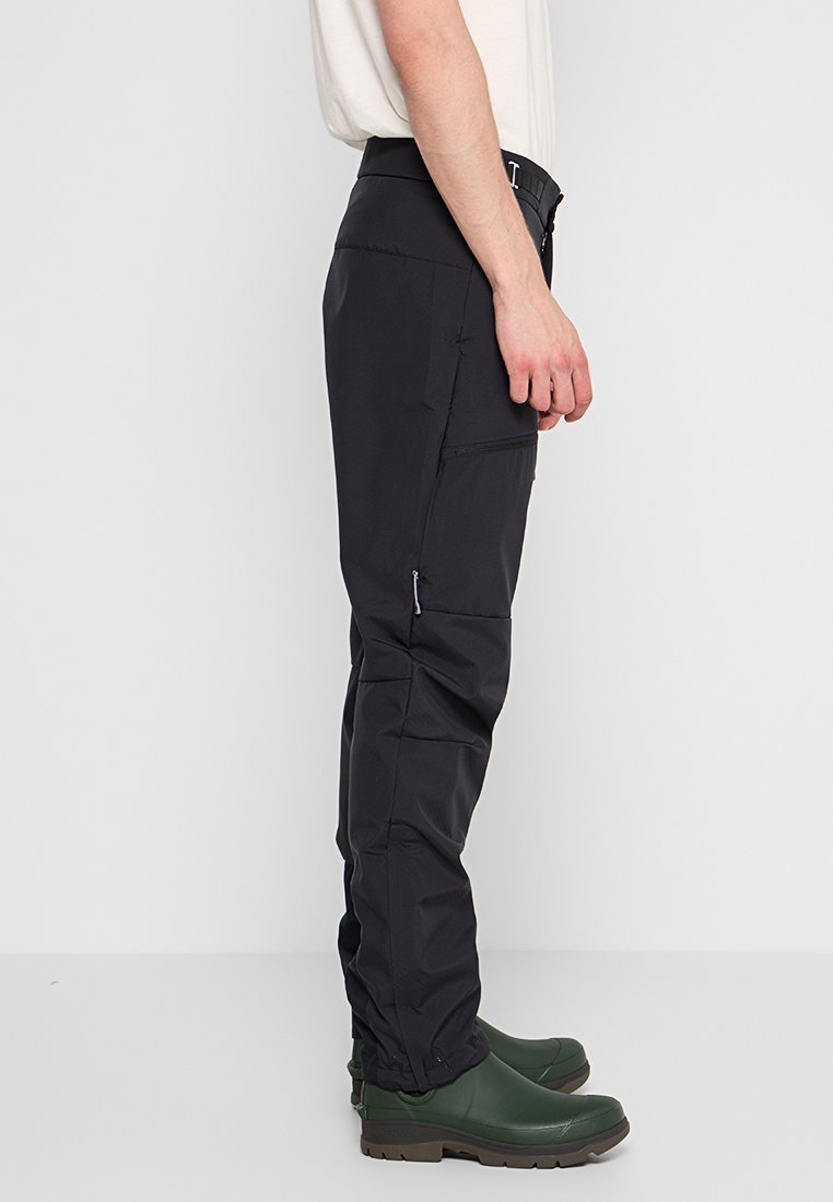 Schwarze wasserdichte Herren-Hosen mit seitlichen Reißverschlusstaschen, elastischem Bund, schmal zulaufendem Schnitt und strukturiertem Stoff. Getragen mit grünen Gummistiefeln.