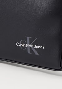 Crna torba od umjetne kože s teksturiranim završetkom, s utisnutim logotipom "Calvin Klein Jeans" u bijelim i sivim odsjajima.