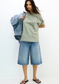 Un t-shirt vert clair à manches courtes associé à un short en denim bleu et une veste en denim. Des tongs complètent la tenue.