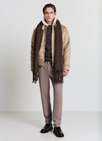 Giacca bomber beige con colletto in pelliccia, maglione scuro, sciarpa marrone con frange, pantaloni marrone chiaro e stivaletti neri.