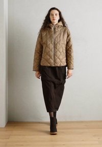 Veste matelassée beige avec une capuche et un motif en losanges, associée à une longue jupe noire et des bottines, se tenant devant un fond neutre.