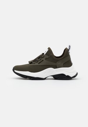 Mørkegrøn strikket sneaker med tyk hvid sål, sort ydersål, elastiske snørebånd og trækflig på hælen på neutral baggrund.