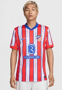 Tricou de fotbal cu dungi roșii și albastre, cu mâneci scurte, design cu guler în V, având un logo și un sponsor imprimate pe față.