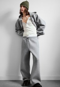 Personne portant un bonnet en tricot vert, une chemise henley blanche, un sweat à capuche gris surdimensionné avec fermeture éclair, un pantalon de survêtement large gris et des chaussures noires, debout à l'intérieur.