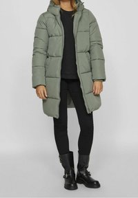 Parka verde acolchada con capucha, cierre de cremallera, ajuste holgado y puños acanalados. Llevada sobre una camiseta negra y combinada con pantalones y botas negras.