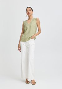 Top senza maniche verde oliva con scollatura a V e dettagli in pizzo. Abbinato a pantaloni bianchi larghi e sandali verdi. Design semplice e casual.