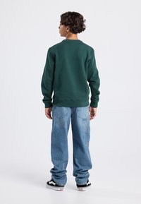 Vihreä huppari ribbipäärmeillä, yhdistettynä vaaleansinihin löysiin farkkuihin, joissa on sauman yksityiskohtia ja sivutaskut, ja päällä mustat lenkkarit.