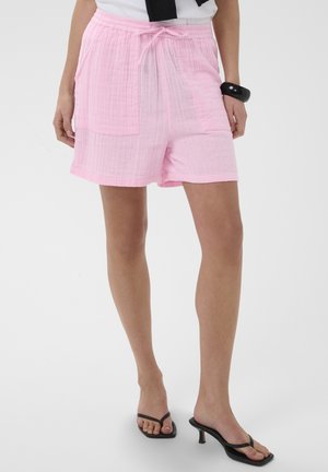 Vrouw draagt loszittende, lichtroze shorts met trekkoord en zakken, gecombineerd met zwarte sandalen met hak en een wit shirt met een zwart accessoire.