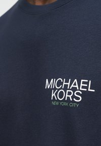 Μπλε σκούρο πουκάμισο με λευκό κείμενο «MICHAEL KORS» και μικρότερο πράσινο κείμενο «NEW YORK CITY» τυπωμένα στο πάνω μέρος του στήθους.